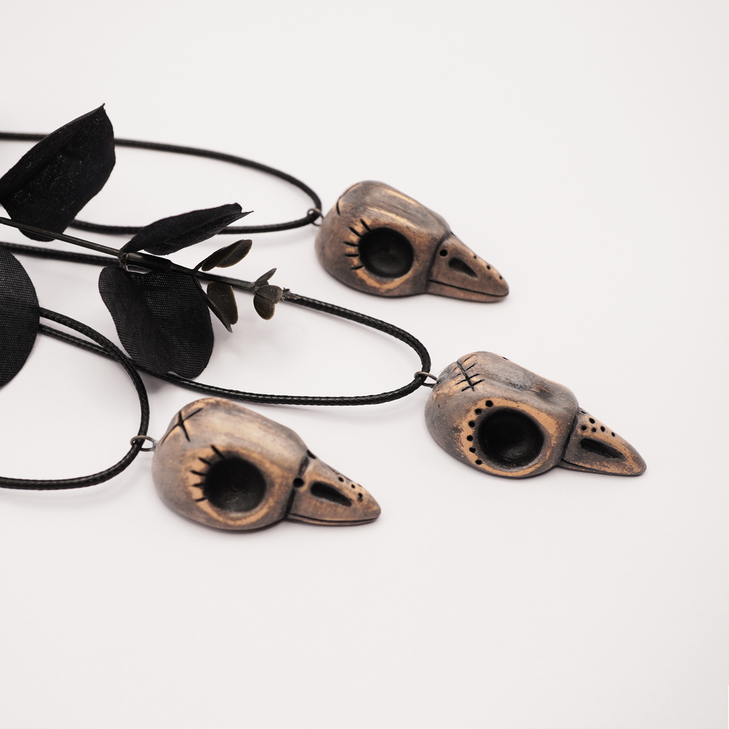 Three handmade ceramic raven skull pendants on black cords displayed on a white background with black decorative leaves.
