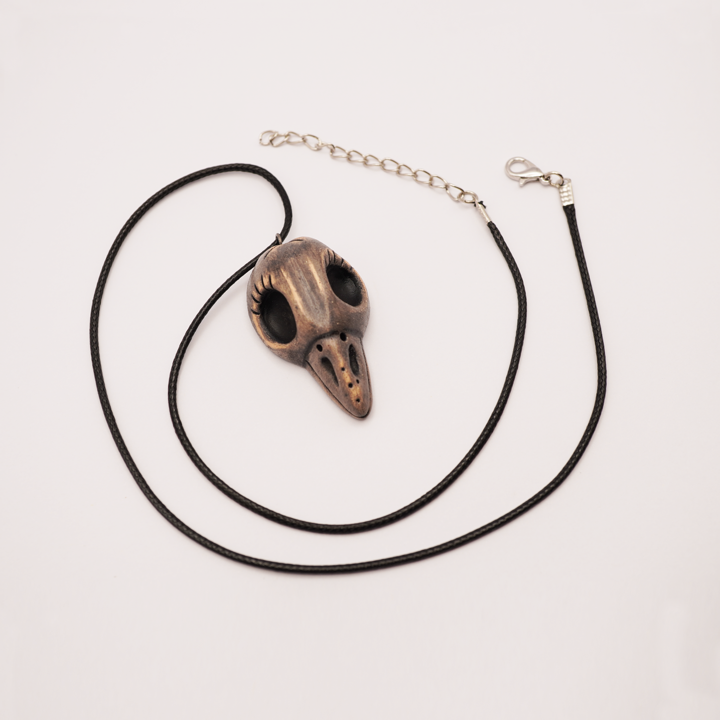 Handmade ceramic raven skull necklace on black cord, displayed on light background.