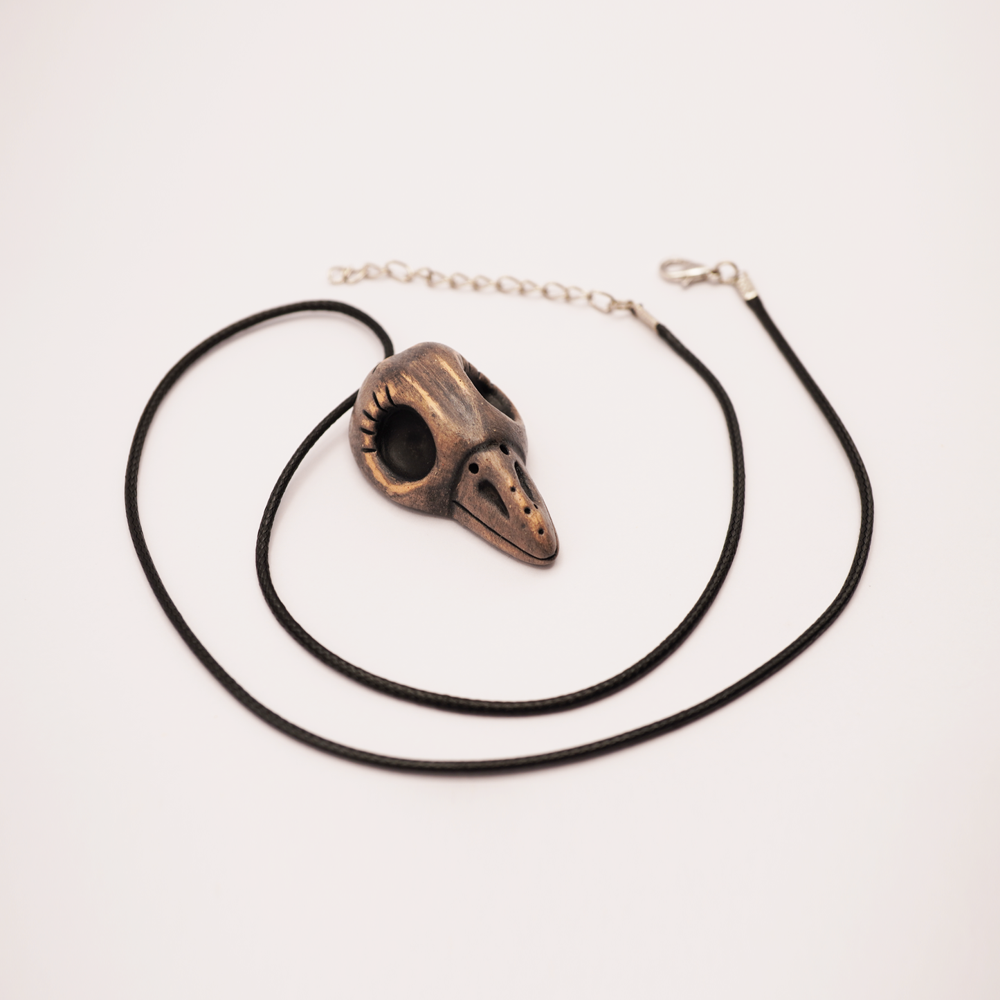 Handmade ceramic raven skull pendant on black cord against white background.