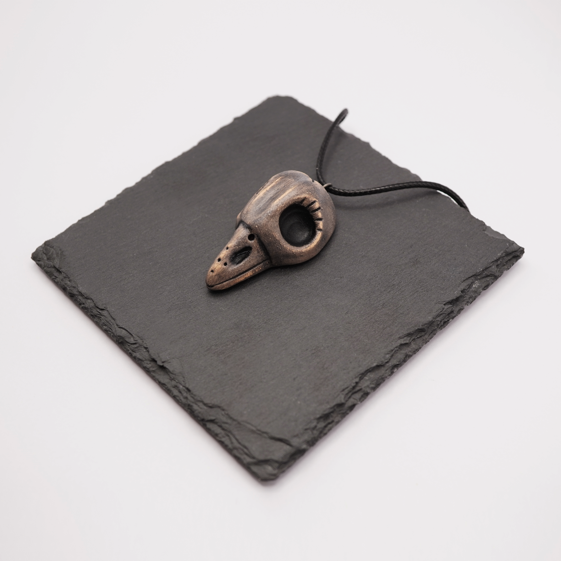 Handmade ceramic raven skull pendant on a black slate display.