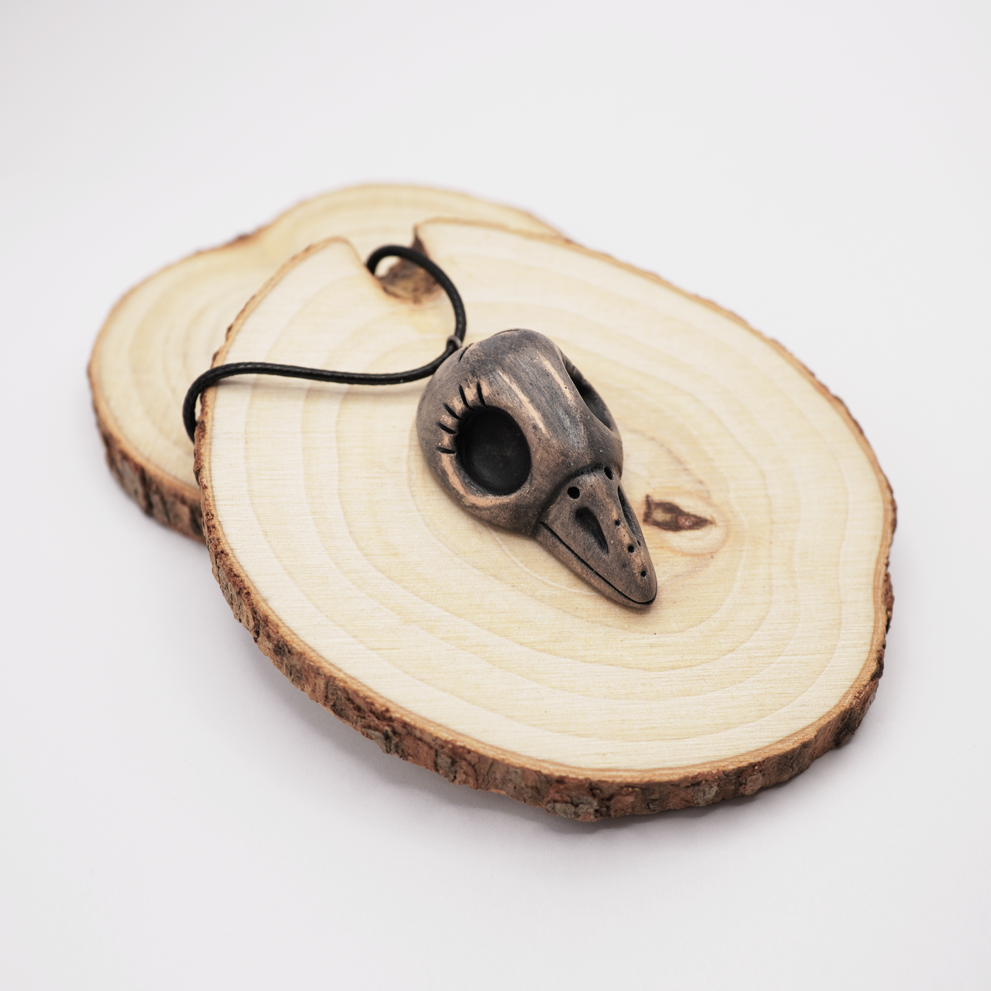 Handmade ceramic raven skull pendant displayed on natural wooden slices.